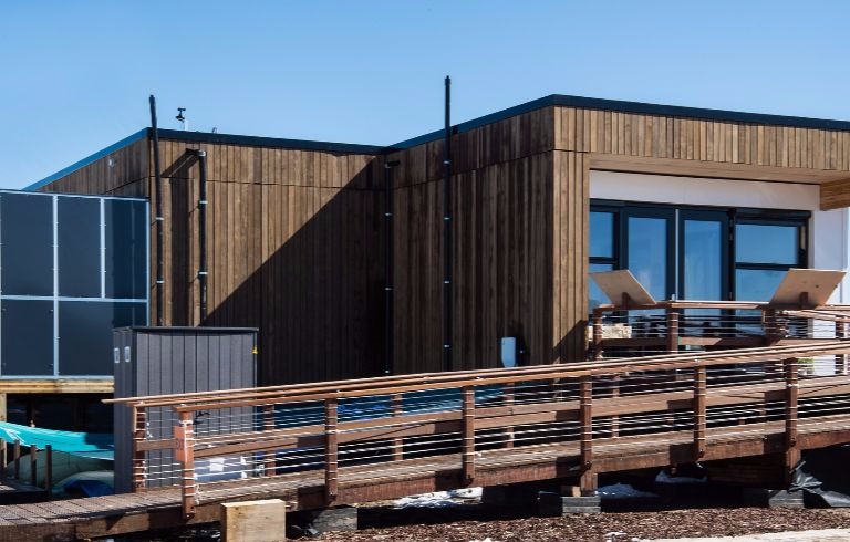 Photo of the exterior of a brown, wood-sided house with a ramp approaching a side entrance with large windows and doors. A dark-colored metal room housing mechanical equipment is visible on the left side. This is the Selficient house by the U.S. Department of Energy Solar Decathlon 2017 team from the HU University of Applied Science, Utrecht, Netherlands. 