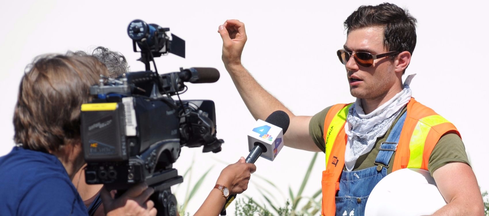 Photo of a young man wearing construction clothing, safety glasses, and carrying a hard hat under his arm, speaking to a TV reporter holding a microphone and a cameraman operating a large camera on a tripod. The young man is a competitor on Team Orange County in the U.S. Department of Energy Solar Decathlon 2015.