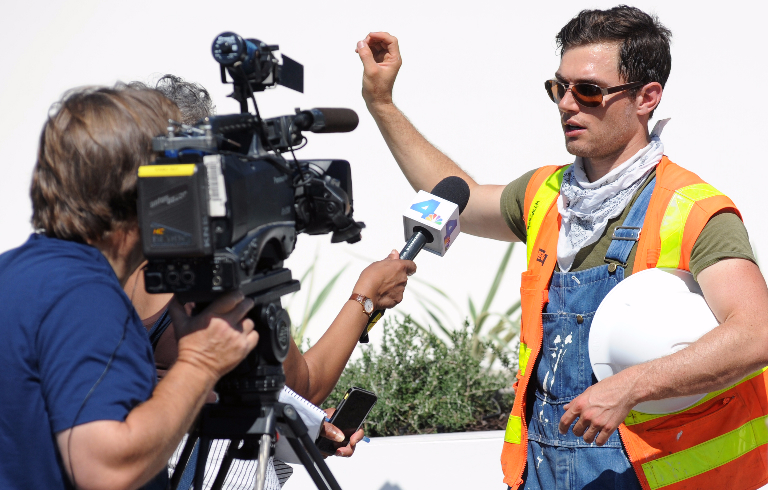 Photo of a young man wearing construction clothing, safety glasses, and carrying a hard hat under his arm, speaking to a TV reporter holding a microphone and a cameraman operating a large camera on a tripod. The young man is a competitor on Team Orange County in the U.S. Department of Energy Solar Decathlon 2015.