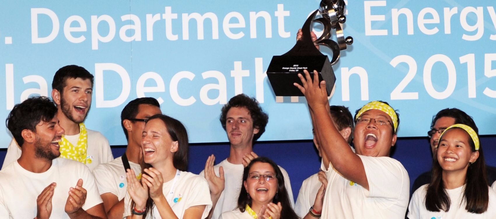 Photo of a group of young men and women wearing white t-shirts, standing in front of a stage back drop. One man is holding a large trophy overhead. This is the U.S. Department of Energy Solar Decathlon 2015 team from Stevens Institute of Technology celebrating its first-place win.