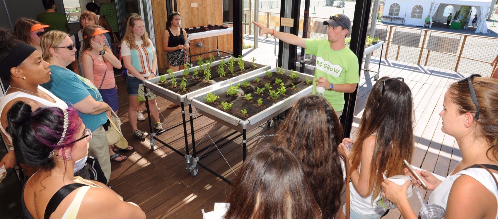 Photo of a young man, a student with the University at Buffalo, U.S. Department of Energy Solar Decathlon 2015 team, providing a tour of the solarium, a glass walled and roofed sun room, of his teams's competition house to a group of visitors. He is gesturing with his hand next to two indoor garden beds with vegetable plants.