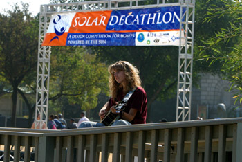 Photo of a woman playing the guitar on a porch, Solar Decathlon sign in background.