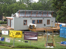 Photo of the rear view of Maryland's Solar Decathlon house, also with a large deck. The back wall has one glass door leading into the house and several smaller windows. The house is under construction and is surrounded by a chain-link fence.