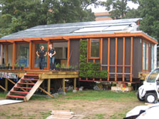 Photo of the front view of Maryland's Solar Decathlon house. The slanted roof is covered in solar panels and the wall off the deck is made of four glass panels. Two students are standing on the deck, surrounded by small potted plants.