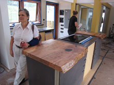 Photo of the kitchen under construction in Maryland's Solar Decathlon house. The island has a stovetop and a counter made of a heavy slab of wood. Two women stand in the kitchen.
