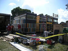 Photo of a house consisting of several modules with sloping roofs and clerestory windows. The house siding consists of multi-color stripes. A partially emptied truck is parked next to the house, and three young men are walking towards the truck. In the foreground is piles of wood, tape marked caution, scaffolding, a ladder, and other construction clutter.