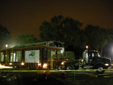 Photo of partially built house mounted on a tractor trailer, lit brightly by several surrounding lights. Trees in the background are also lit up against a dimly lit night sky. The rear end of the house features large glass windows, through which the interior of the house can be seen.