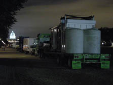 Photo taken at night of several large trucks standing in line, with a cargo of house modules on board each. The truck drivers are awaiting a signal that the time has come to drive onto the Solar Decathlon site on the National Mall. The well-lit Capitol is prominent in the background.