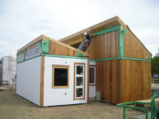 Photo of a house with a larger rear portion and a smaller front portion, both of which are rectangular with a roof sloped toward the south. A student wearing protective gear stands on the roof of the front portion and secures the roof of the rear portion in place.