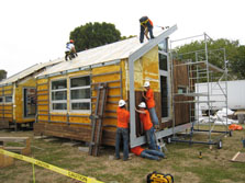 Two people wearing safety gear attach solar panels to the roof of a house, while three people install dark wooden siding to the walls, covering up a rough-looking layer of insulation.