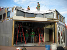 Photo of a home with a large open gap spanning one corner. Two people, several ladders, and interior trim are visible through the gap. Two other people are standing on the roof.