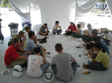Photo of a group of 16 students sitting in a circle on the floor in the corner of a large white tent, eating food off of paper plates and drinking out of foam cups.
