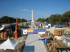 Aerial photo of the National Mall features a square metal entrance gate with a banner that says Solar Decathlon, with a walkway extending into the distance to a large tent. On both sides of the walkway are solar houses that appear nearly complete.