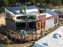 Aerial photo of a house that is nearly fully assembled. Three student are installing solar panels on the roof while two install railings on the deck.