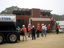 Photo of a water tanker truck with a hose leading to the solar house. Eight people are lined up holding the hose, with one man on the top of the shed containing a storage tank.