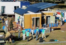 Photo of a home under construction. The home has several large solar panels on the roof, and people are walking around the home and working on it.