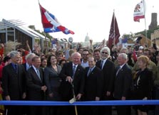 Photo of Secretary Bodman cutting a wide ribbon with a pair of oversized scissors. He is flanked by several men and women in business suits with a crowd of college students behind him and the Capitol Building visible in the background.