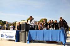 Photo of a cluster of people standing behind a long, low table, with a young man in a black T-shirt speaking from a podium.