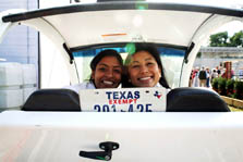 Photo of the faces of two smiling young woman with cheeks touching. They are sitting in the front seat of a very small white car.