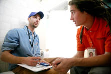 Photo of two men sitting and talking to each other. The one on the left is in a long-sleeved blue denim shirt and wearing a baseball cap; the one on the right is wearing an orange polo shirt.