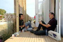 Photo of two young men sitting facing each other on the passageway of the deck of a house. They are eating sandwiches.