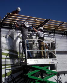 Photo showing two men in hard hats standing on a platform sitting atop a scissors lift, reaching up and taking apart a wooden lattice-work on the roof of the solar home.