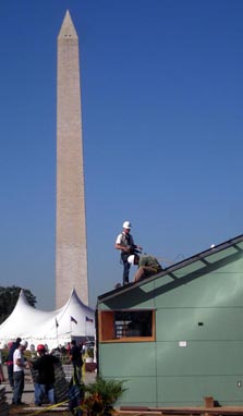 Photo of a solar house, with two men in hard hats working on the roof; the Washington Monument is in the background.