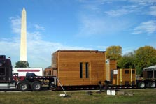 Photo of a tractor-trailer with a square unit of a solar house sitting on the trailer.