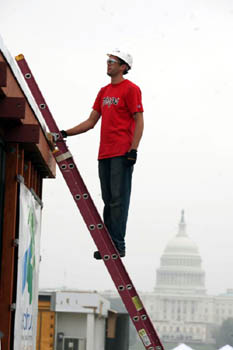 Photo of a young man on a ladder that is leaning against a house. The Capitol building is visible in the background.