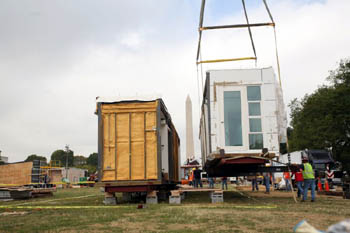 Photo of a crane holding half of a house suspended several feet and to the right of several footings that will serve as its foundation. The other half of the house is already in place. The Washington Monument is visible in the background between the two halves of the house.