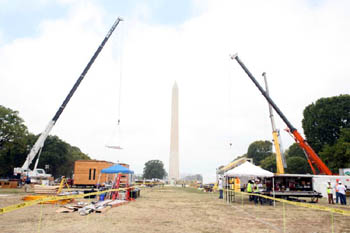 Photo of a dirt path extending toward the obelisk of the Washington Monument. One large crane is extended to the left of the path, while two are extended to the right. Homes in various states of construction line the path.