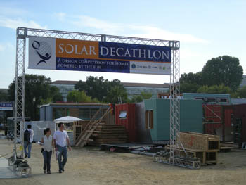 Photo of people walking through a metal gate with sides about a foot square and about 15 feet high, a top about a foot square and about 20 feet wide, and a Solar Decathlon banner across the top. A jumble of buildings is in the background.