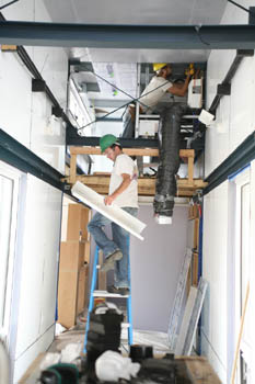 Photo of the interior of a narrow building, where one young man works on equipment in the ceiling and another carries a piece of metal while climbing a ladder.