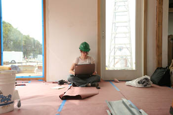 Photo of a young woman in a hardhat leaning against a wall with her legs crossed, a laptop computer in her lap. The surrounding room shows evidence of ongoing construction, and a ladder is visible through an adjacent window.