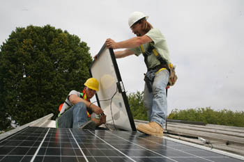 On a sloping rooftop and wearing safety equipment, a young man sits and wires a solar panel while an older man holds the solar panel in place. In the foreground is a solar panel that has already been installed.