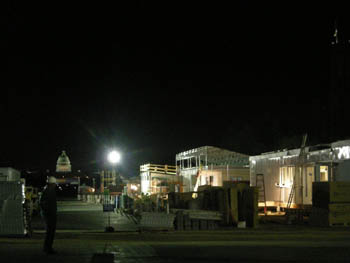 Photo looking east from the center of the Solar Decathlon shows a man in a hardhat on a walkway, looking down a row of houses lit by a temporary light.