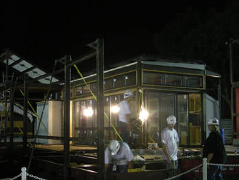 Photo of four people working in the midst of a network of girders laid out to form a deck in front of a house with an exterior made mostly of glass. At the front of the deck, a number of girders form a vertical structure.
