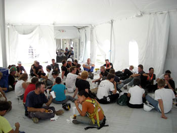 Photo of a large group of people sitting on the floor in the corner of a large white tent, eating food and drinking.