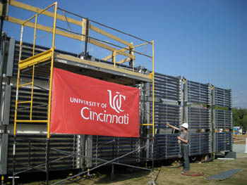 Photo of a young man cleaning a large bank of glass tubes that form the entire year wall of the a house. A scaffold in front of one section of glass tubes carries a banner that says 'University of Cincinnati.'