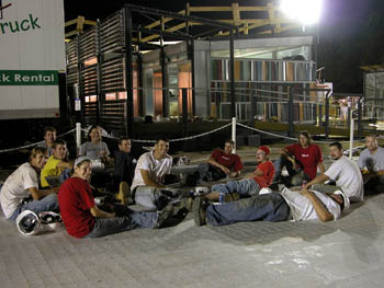 Night photo of 13 young men and women sitting, reclining, and laying down on walkway tiles in front of a house lit by bright portable lights.