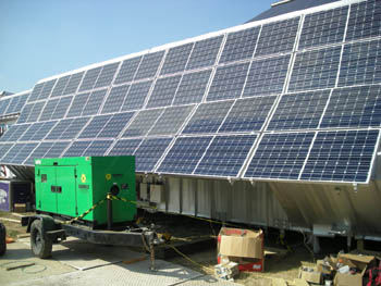 Photo of a green electrical generator in the foreground, with a large array of photovoltaic panels in the background.