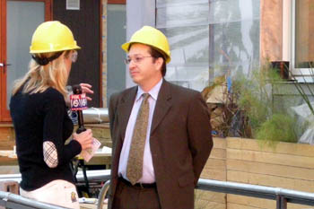 Photo of a woman reporter holding a microphone and interviewing a man dressed in a suit and wearing a hard hat.