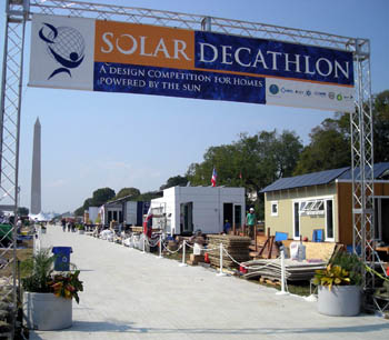 Photo showing the exteriors of four solar houses lining a gravel path. The Solar Decathlon banner arches over the pathway in the foreground, and the Washington Monument is visible in the distance.
