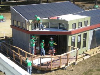 Aerial photo of the front porch of a solar house under construction, with four men in green shirts painting the walls and railing.