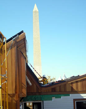 Photo of a worker in a hard hat tethered to the roof and working on skylights. The Washington Monument looms in the background.