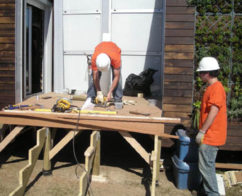 Photo of the exterior of a solar house under construction, with two men in hard hats building the framework for a front porch. Behind one of the men the side of the house is visible and contains panels of growing plants.