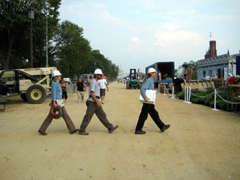 Photo of three people in hardhats crossing a dirt path in single file, with solar houses under construction in the background.