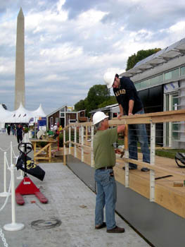Photo of two men in hardhats working on the railing of a ramp, with the solar home in the background.