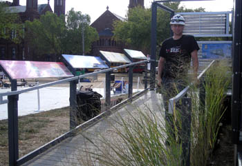 Photo of a man standing at the foot of a ramp leading into a solar house, with signs placed along the railing.