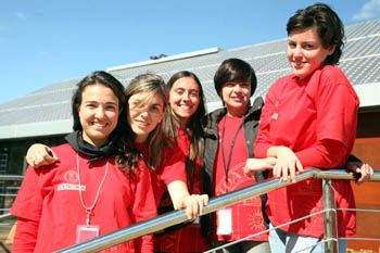 Photo of five women in red T-shirts leaning on a rail and standing in front of a house with solar panels on top.